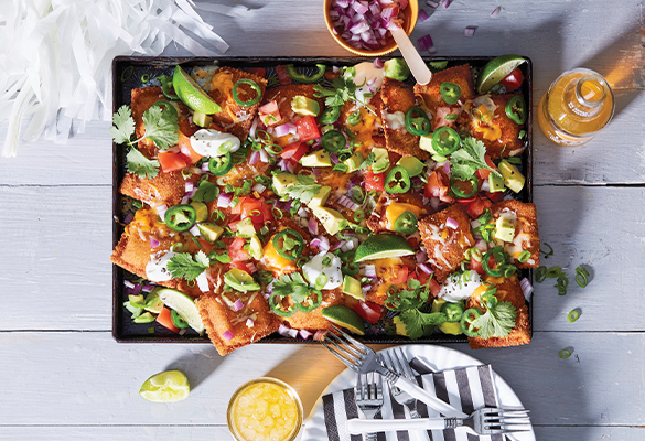 Photo showing a top-down look at Golden Raviolio Nachos in a black plate on a white wooden table backdrop surrounded by a white pom-pom, yellow bowl of diced onions, orange drink, fresh lime wedge and a plate and fork.