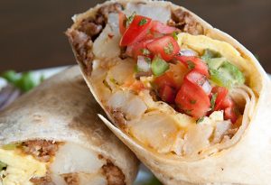 Photo close up of a burrito cut in two and stacked on each other. Burrito is filled with meat, cheese, potatoes, onions and tomatoes.