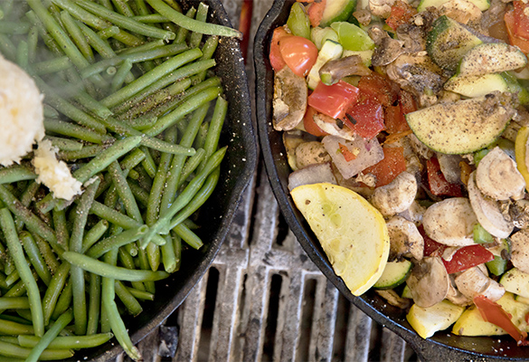 Photo of two cast iron skillets on a grill filled with veggies. Left skillet is filled with green beans, the right skillet is a mix of tomatoes, peppers, onions, squash and mushrooms.