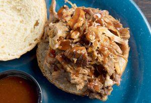 Photo of an open pulled pork sandwich with a drizzle of BBQ sauce on a blue plate with a small, round container of BBQ sauce.