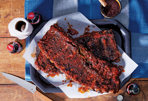 Photo of a rack of ribs with sauce on a wood table and a blue checkered cloth. Bottles of coke, a container of sauce, a roll of napkins and a knife surround the platter of ribs.