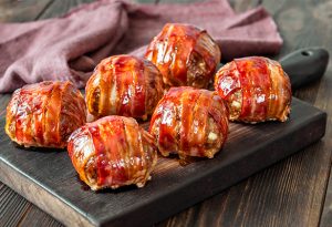 Photo of six bacon wrapped meatballs on a wooden board.