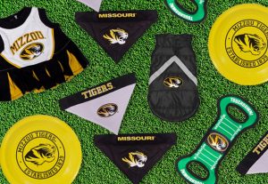 Composite graphic of different Mizzou branded pet products on a green grass background including pet cheerleading uniform, bandanas, puffer vest, chew toy, and frisbee