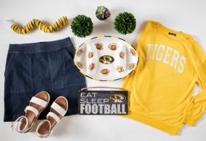 Outfit of the Week showing a photo flat lay of a tiger tail, denim skirt, white platform sandals, ceramic food tray, "Eat Sleep Football" wall sign and a gold "Tigers" sweater.